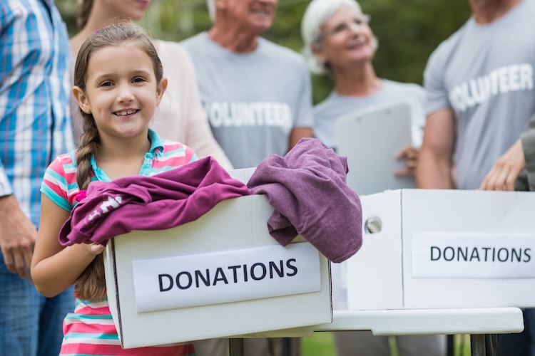 A young girl with a braid and a striped shirt is smiling as she places clothing into a box labeled 'DONATIONS'. Behind her, a group of cheerful adults wearing 'VOLUNTEER' t-shirts are gathered, likely part of a community donation event outdoors.