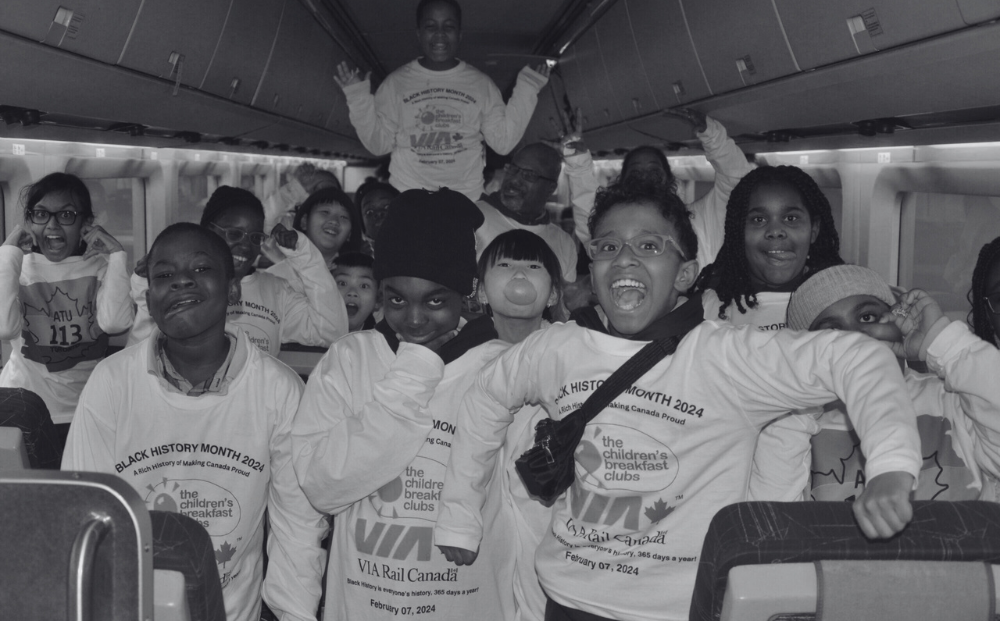 A group of cheerful children wearing matching shirts posing for a photo on a train, celebrating an event, with one child standing and others sitting, some making playful gestures, reflecting a joyous and energetic atmosphere.