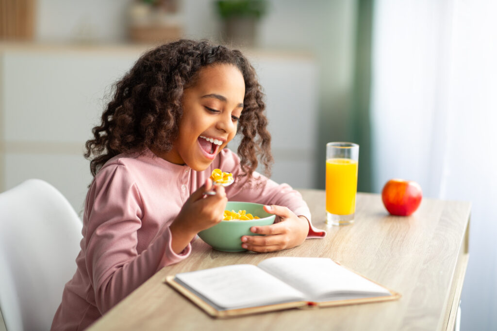 A cheerful young girl enjoys her breakfast while reading from an open book on the table, highlighting the importance of a nutritious morning meal in supporting academic success and concentration in school.