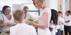 A friendly volunteer serving breakfast to a line of eager children, with a backdrop of a busy, vibrant kitchen. 